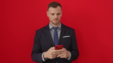 Rahat görünümlü, akıllı telefon tutan, doğal ve basit yüzünde ciddi bir ifade sergileyen, kırmızı arka planda izole edilmiş genç İspanyol bir adam, kameraya bakıyor.