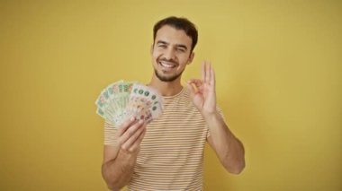 Neşeli genç İspanyol bir adam bir avuç dolusu para tutuyor, bir parmak hareketiyle mükemmel bir işaret veriyor, izole edilmiş sarı arka planda serinleyerek gülümsüyor..