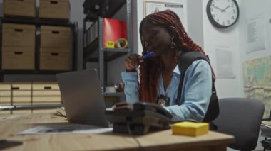 Afro-Amerikalı bir kadın dedektif bir ofis ortamında dizüstü bilgisayar ve dosyalarla kanıtları analiz ediyor..