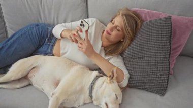 Evcil köpeğiyle kanepede uzanmış, akıllı telefonunu evde kullanırken gülümseyen genç bir kadın..