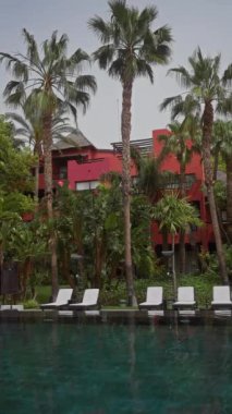 Palmiye ağaçlarıyla çevrili bir havuzu olan sakin bir tropikal tatil köyü ve kırmızı bir binaya karşı güneş pansiyonları..