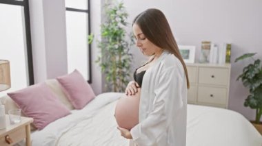 Sakin, genç, hamile, İspanyol bir kadın düşünceli bir şekilde yatak odası düzeninde duruyor, annelik beklentisinin somutlaşmış hali..