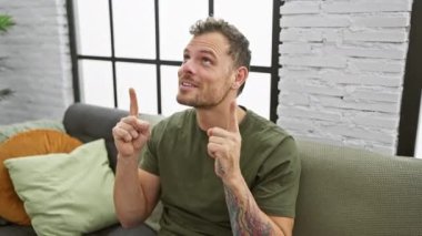 Şaşırmış genç adam sevinçle evinin kanepesinde otururken yukarı doğru işaret ediyor. Şaşırma ve şaşkınlığın portresi yakışıklı yüzünü süslüyor..