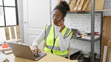 Atölyede bilgisayar ve telefon kullanan güvenlik yeleği giyen Afrikalı Amerikalı kadın.