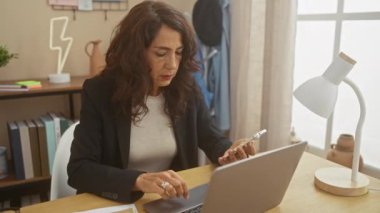 Modern bir ofis ortamında akıllı telefon ve laptop kullanan orta yaşlı bir kadın..
