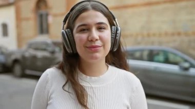 Genç bir İspanyol kadın, bir şehir caddesinde durup, şehir yaşam tarzı ve rahatlık hissini aktarırken kulaklıkla müzik dinlemekten hoşlanıyor..
