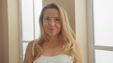 Elinde makyaj fırçası olan gülümseyen sarışın bir kadın, beyazlar içinde, güneşli bir dairede poz veriyor.