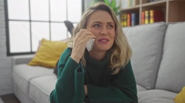 Gülümseyen beyaz bir kadın, rahat, aydınlık bir oturma odasında telefonla konuşuyor, rahat ve zarafet saçıyor..