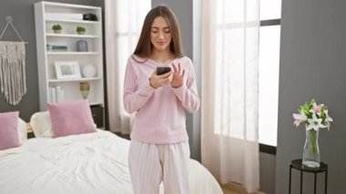 Pijamalı, gülümseyen kadın modern dekorlu, parlak yatak odasında akıllı telefon kullanıyor.