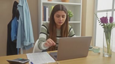 Pencere kenarındaki masada dizüstü bilgisayar, gözlük ve hesap makinesiyle çalışan İspanyol kadın.