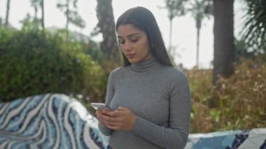 Genç bir İspanyol kadın akıllı telefonunu palmiye ağaçlarıyla dolu bir parkta kontrol ediyor..