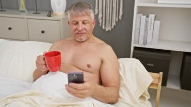 Orta yaşlı, yatakta gri saçlı, elinde kırmızı bir kupa ve akıllı bir telefon tutan, rahatlama ve modern ev içini gösteren bir adam..