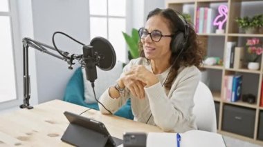 Gülümseyen İspanyol kadın modern stüdyoda mikrofon ve tabletle podcast yapıyor..