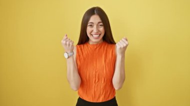 Heyecanlı ve gururlu bir zafer kutlaması! Güzel İspanyol kadın, kolsuz tişört giyiyor, kollarını kaldırıp bağırıyor. Sarı izole stüdyo arka planında neşeli bir başarı.