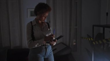 Afro-Amerikalı bir kadın evdeki suç mahallini araştırıyor. Akıllı telefon ve kalemle notlar alıyor. Mavi ve kırmızı ışıklarla aydınlatılıyor..