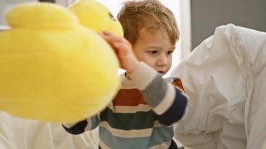 Sarışın bir çocuk, evdeki parlak, rahat yatak odasında bir yatakta otururken sarı bir topla oynuyor..