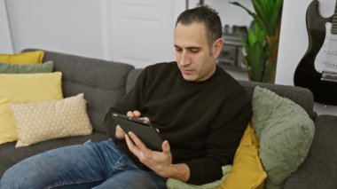 Gülümseyen bir adam rahat bir şekilde gri bir koltuğa tabletle oturur ve rahat bir oturma odasında renkli yastıklarla süslenir..