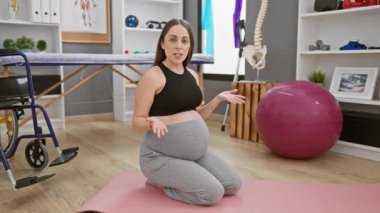 Aktif kıyafetli hamile bir kadın fizik tedavi kliniğinde yoga minderinde oturuyor..