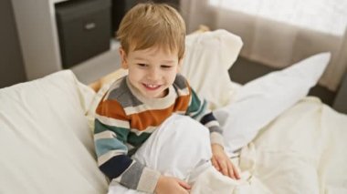 Evde güneşli bir yatak odasında beyaz bir yatakta oturan çizgili süveterli, gülümseyen sarışın çocuk..