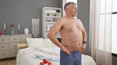 Beyaz saçlı orta yaşlı bir adam yatak odasında dikiliyor, dalgın görünüyor ve pijama giyiyor..