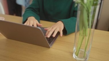 Çiçeklerle çevrili ve yeşil bir kazak giyen, dizüstü bilgisayarda çalışan, sarı saçlı, gülümseyen genç bir kadın..