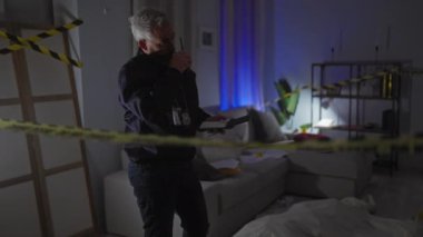 Sakallı ciddi bir adam, kapalı bir suç mahallinde telsiz kullanıyor.