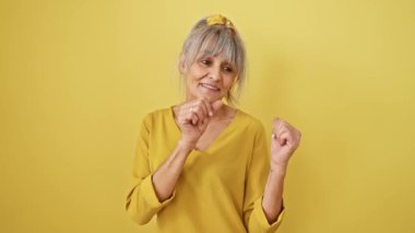 Beyaz saçlı, neşeli at kuyruklu, kendinden emin orta yaşlı bir kadın. Başparmaklarının arkasını göstererek mutlu bir şekilde izole edilmiş sarı bir arka planda ışıldıyor..