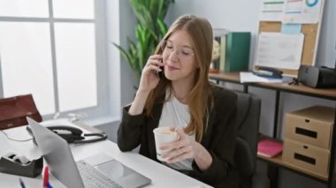 Gözlüklü profesyonel bir genç kadın, telefonda konuşuyor ve iyi aydınlatılmış bir ofiste kahve fincanı tutuyor.