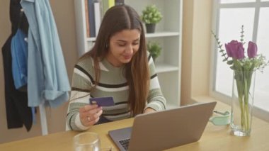 Genç İspanyol bir kadın evde online alışveriş için kredi kartı kullanıyor. Tarz ve konfor yayıyor..