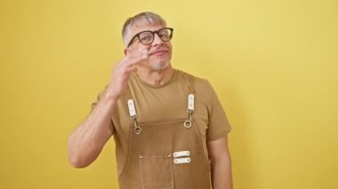 Gri saçlı, kendine güvenen orta yaşlı bir adam, neşeli bir şekilde gülümseyen, sarı arka planda şık gözlükler ve önlükler takan. Elleri saçlarına dokunuyor, biraz da cazibe katıyor..