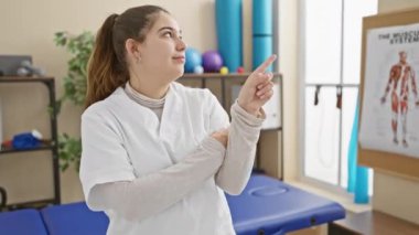 Sıcak bir gülüşü olan neşeli, genç İspanyol hemşire, rehabilitasyon merkezindeki iş yerinde dostça bir jest yaparak, sevinçle kenarı işaret ediyor.