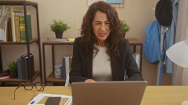 Orta yaşlı bir kadın, evdeki ofis masasında dizüstü bilgisayarla çalışırken, etrafı kitaplar ve bitkilerle çevrili bir şekilde rahatsızlığını dile getiriyor..