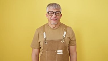 Orta yaşlı, gri saçlı, önlük takan, gözlüklü, gülümseyen, el ve parmaklarıyla telefonla konuşan bir adam. İletişim kavramları. izole edilmiş sarı arkaplan