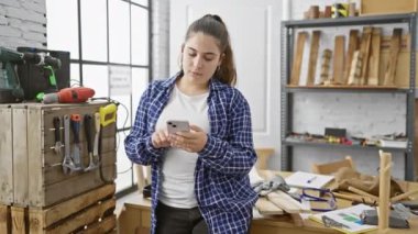 Marangozluk atölyesinde ahşap ve aletlerle akıllı telefon kullanan İspanyol kadın..