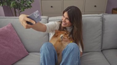 Genç bir kadın Pomeranian köpeğiyle bir selfie çekiyor, rahat bir oturma odasında kanepede sarılıyor..