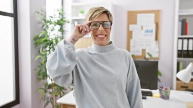 Kısa saçlı ve gözlüklü gülümseyen profesyonel bir kadın ofis ortamında kendine güvenerek pozitiflik ve işyeri rahatlığı tasvir ediyor..