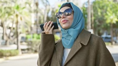 Güneşli şehir sokağında akıllı telefon kullanan tesettürlü olgun kadın, bağlantı ve çeşitliliği yansıtıyor.