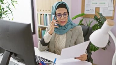 Tesettürlü profesyonel bir kadın ofiste çalışıyor, bir belgeyi okurken telefonla konuşuyor..