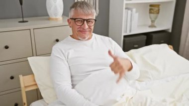 Orta yaşlı, gri saçlı, pijama giyen, gülümseyen, el ve parmaklarıyla telefonla konuşan bir adam. İletişim kavramları. yatak odasında