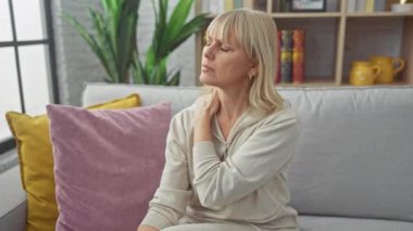 Düşünceli sarışın bir kadın rahat bir oturma odasında kanepede otururken boynu ağrır..