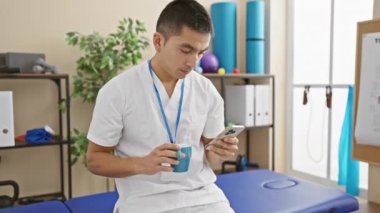 Klinikte elinde kahve fincanı ve akıllı telefonla gülümseyen İspanyol erkek hemşire..