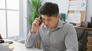 Ofisteki genç Asyalı bir adam, modern iş ortamının ortasında akıllı, odaklanmış ve profesyonel bir telefonla konuşuyor..