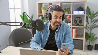 Sakallı, yakışıklı bir İspanyol, içeride bir radyo programı sunuyor. Mikrofon, dizüstü bilgisayar ve akıllı telefon kullanıyor..