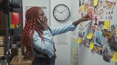 Dedektiflik bürosundaki genç bir Afro-Amerikalı kadın telefonda konuşurken bir tahtadaki kanıtları analiz ediyor, odaklanma ve kararlılığını iletiyor..