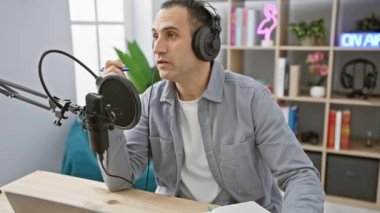 İspanyol adam modern radyo stüdyosunda mikrofona konuşuyor. Renkli bir kitap arkaplanı var.