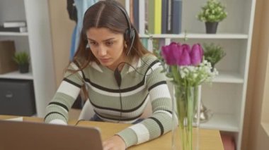 Kulaklığı olan odaklanmış bir İspanyol kadın çiçeklerle donatılmış modern bir ev ofisinde laptopuyla çalışıyor..