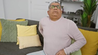 Rahat bir oturma odasında kanepede otururken karın ağrısı çeken gözlüklü olgun bir adam..