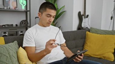 Beyaz tişörtlü, düşünceli bir İspanyol adam modern oturma odasındaki tabletle etkileşime geçiyor..