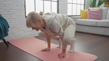 Gündelik kıyafetli sarışın bir kadın modern bir oturma odasında pembe bir paspasla yoga yapıyor..