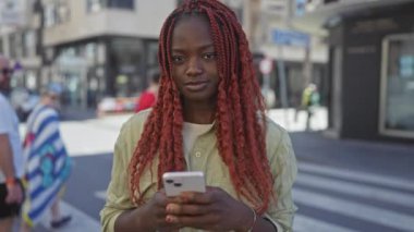Kırmızı örgülü gülümseyen Afro-Amerikan kadın canlı bir şehir caddesinde akıllı telefon kullanıyor.